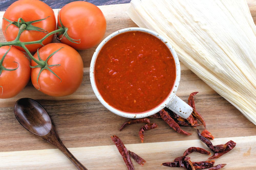 a cup of red tamale sauce surrounded by dried chiles, tomatoes and corn husks