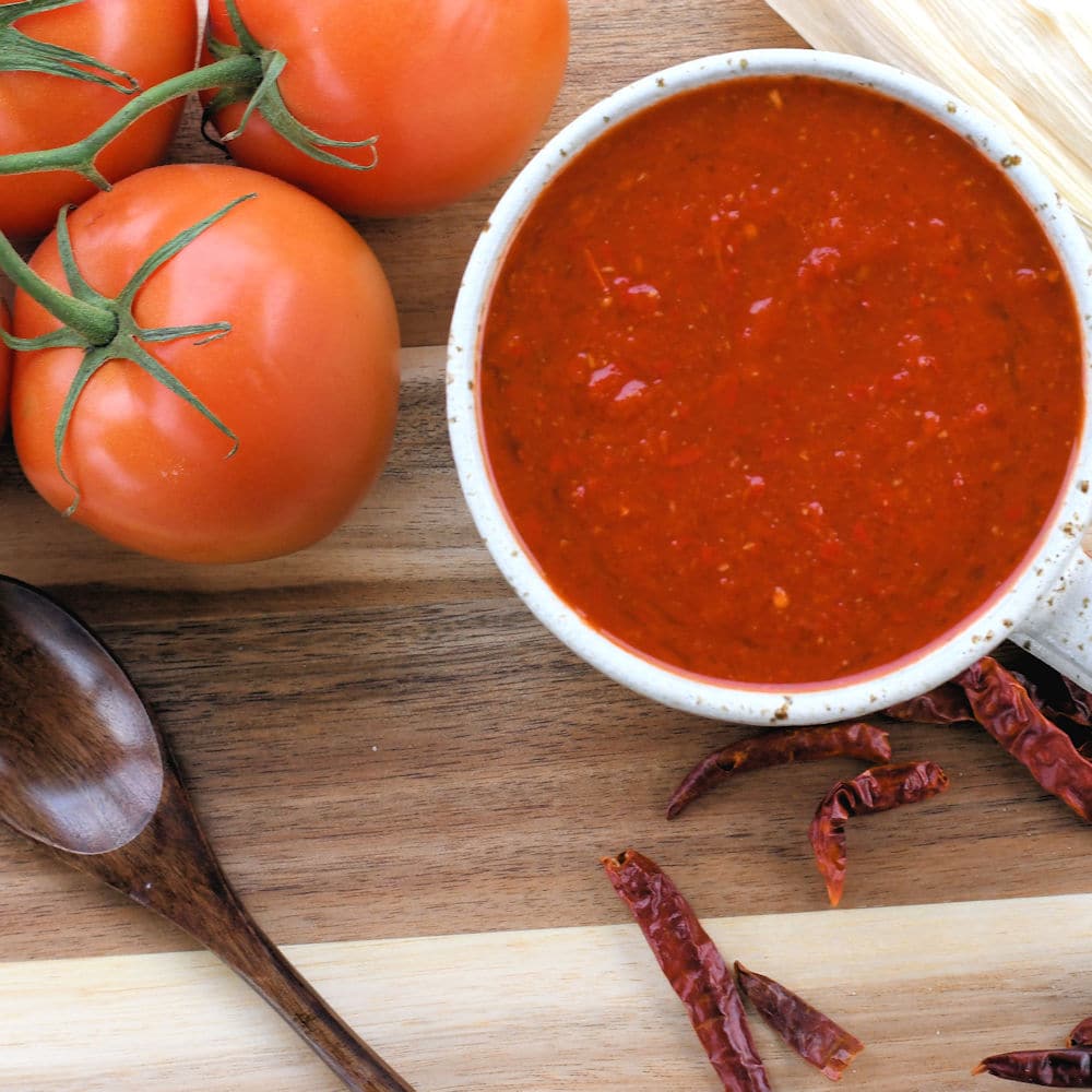 red tamale sauce in a rustic white cup shown on a wooden counter with dried red chiles and tomatoes.