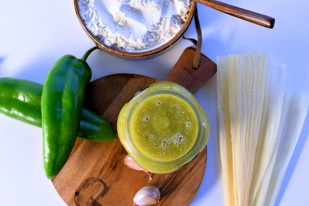 green tamale sauce in a jar shown on a counter with anaheim chiles and corn husks.