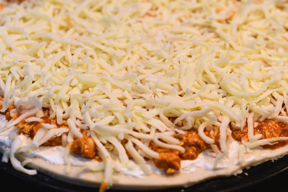 shredded cheese place on top of buffalo chicken pizza, ready for the oven