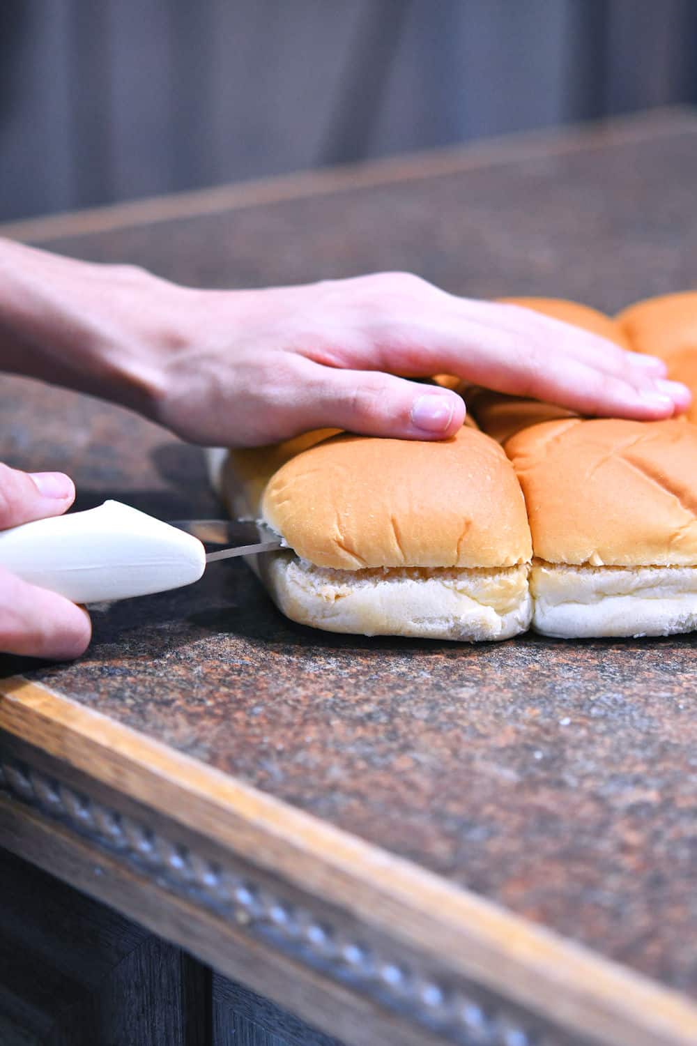 dinner rolls on a counter being cut through the horizontal center with a long serrated bread knife