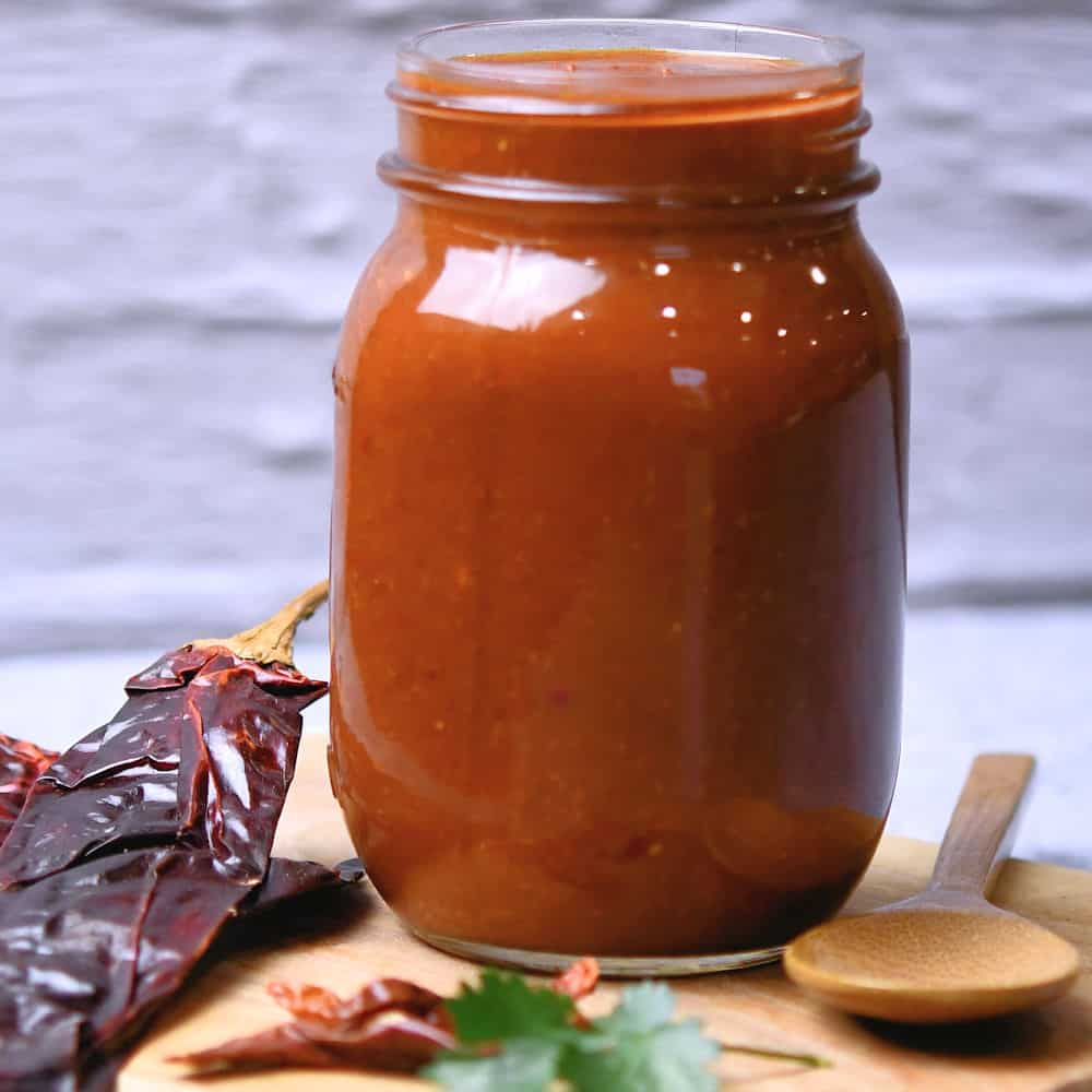 Red enchilada sauce in a glass jar, displayed with dried California chiles on a wooden cutting board.