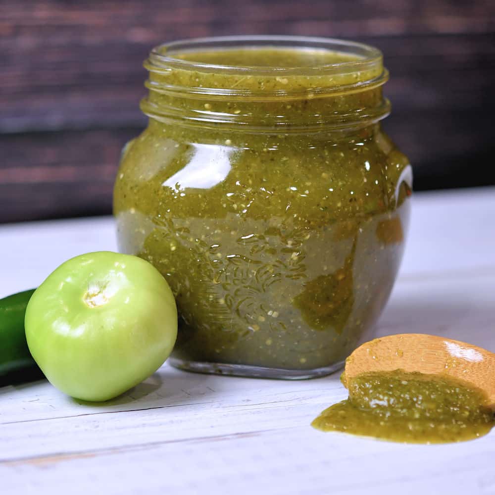 Green enchilada sauce in a glass jar, displayed on a wooden table.