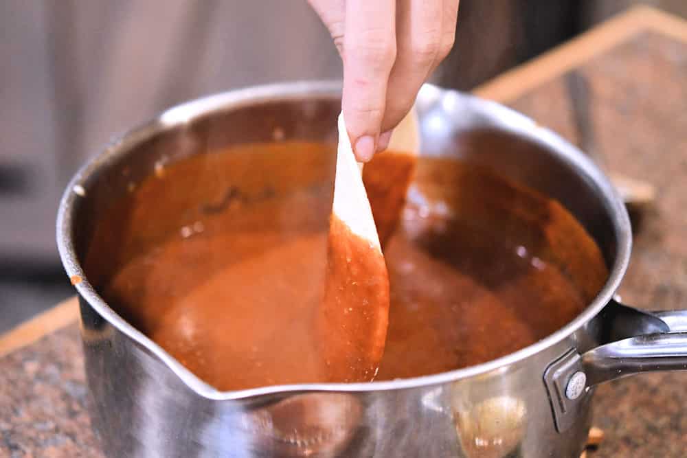 Dipping a corn tortilla into a saucepan of red enchilada sauce.