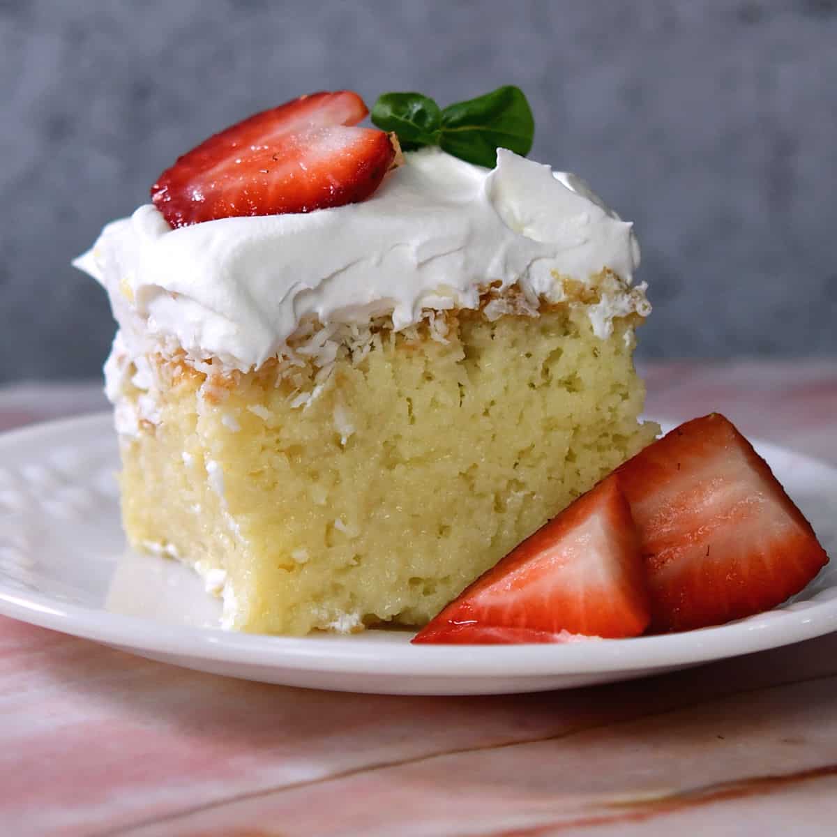 A piece of coconut tres leches cake on a white plate, garnished with strawberries.