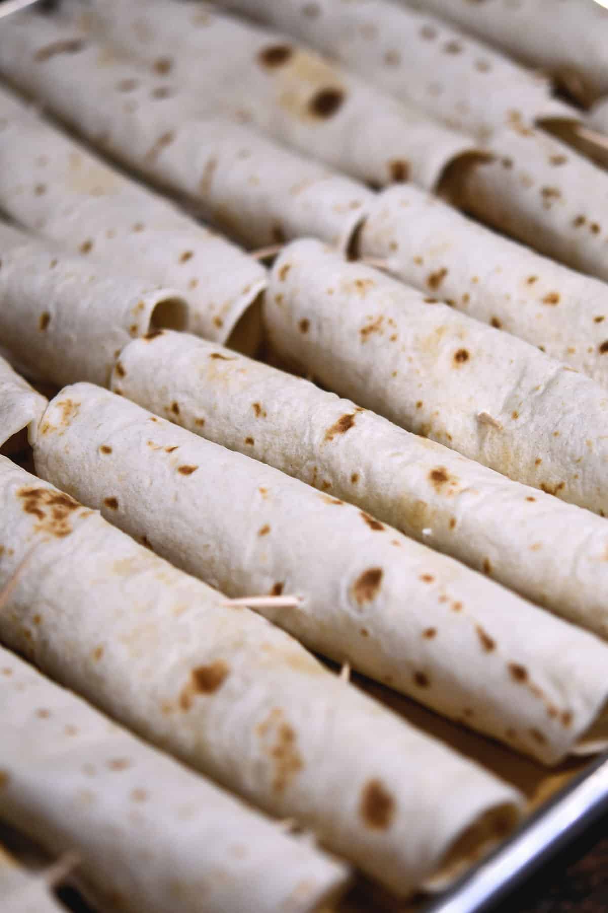 a try of rolled flour tacos, pierced with toothpicks ready to be baked