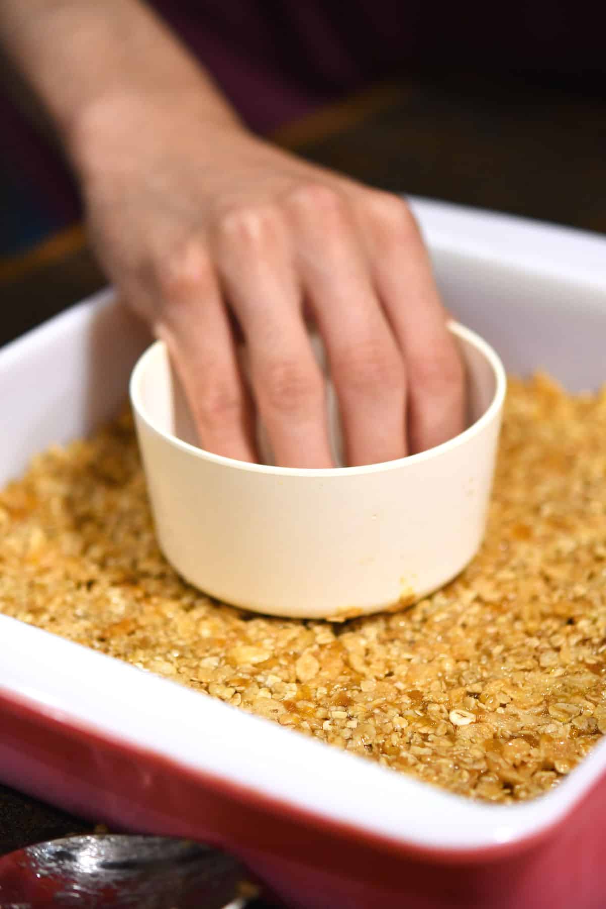 using a measuring cup to pack granola into baking dish