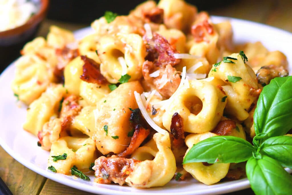 A white plate filled with creamy tortellini pasta garnished with crispy bacon pieces and fresh parsley. The dish is topped with grated cheese and accompanied by a fresh basil sprig on the side.