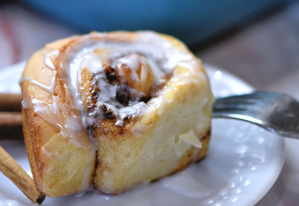 A close-up of a cinnamon roll drizzled with icing, placed on a white plate. The roll's swirls reveal a rich filling of cinnamon and sugar. A fork rests on the side of the plate, and two cinnamon sticks are visible in the background.