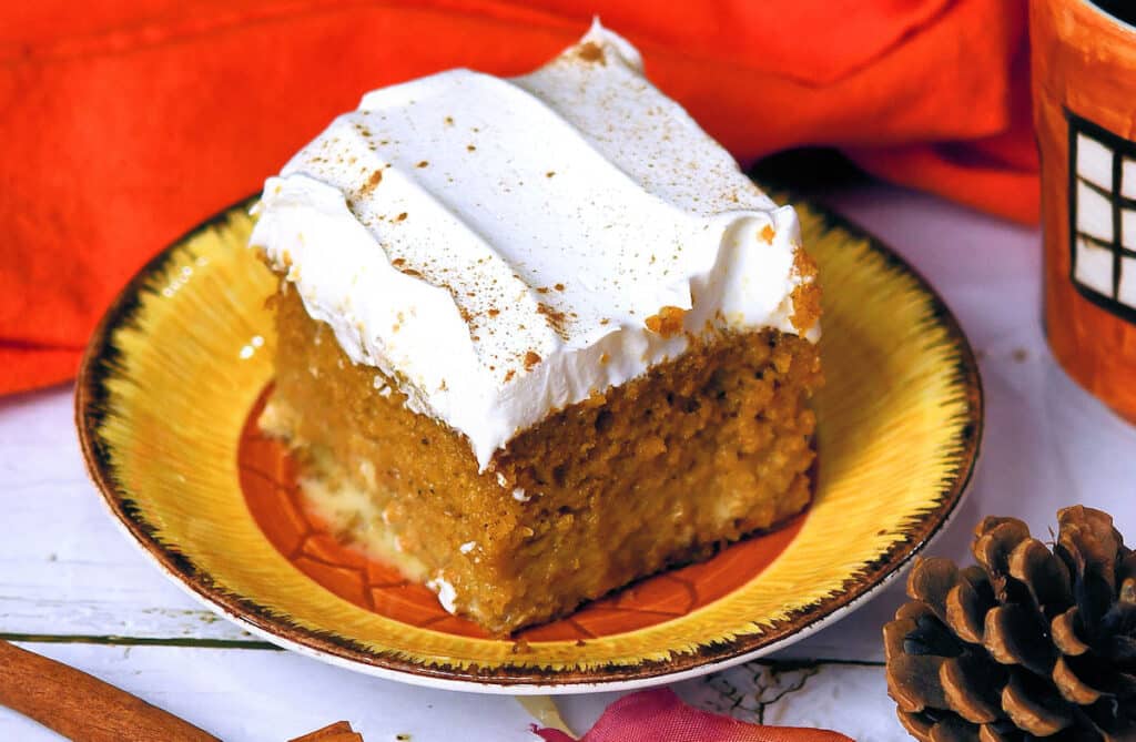 A slice of moist pumpkin cake with a thick layer of white frosting, lightly sprinkled with cinnamon on top. The cake sits on a yellow-orange plate. In the background, there is an orange cloth and a pinecone, suggesting an autumn theme.