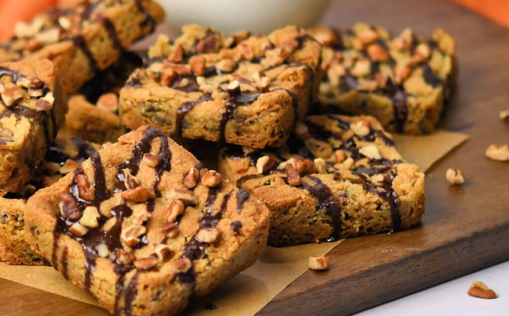 cookie bars on a wooden cutting board, drizzled with chocolate and chopped pecans
